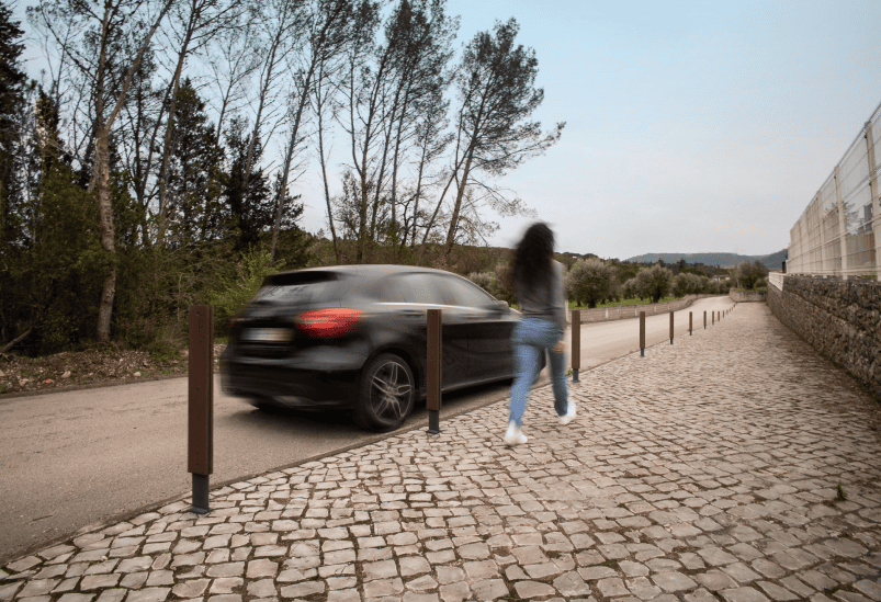 Calle con coche y mujer caminando por la acera con pilonas Floema Frame de plástico reciclado.