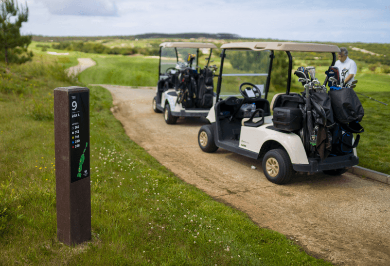 Tee totem Royal en campo de golf para áreas de tee, fabricado en plástico reciclado marrón y HPL+. Son visibles dos buggies de golf con bolsa de palos y un jugador.