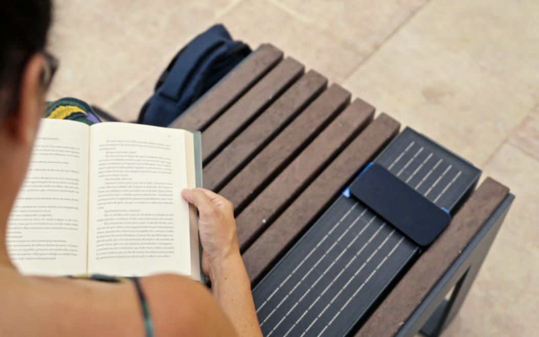 Vista superior de una persona leyendo en un banco inteligente hecho de plástico reciclado, mientras carga un teléfono móvil en el módulo alimentado por energía solar.