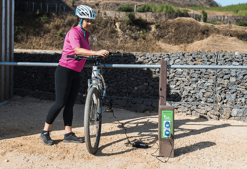 Persona cargando bicicleta eléctrica en conjunto E-bike charger Nature y aparcamiento para bicicletas Triatlo, modelos Floema, con estructura de plástico reciclado, equipamientos para la movilidad eléctrica suave.