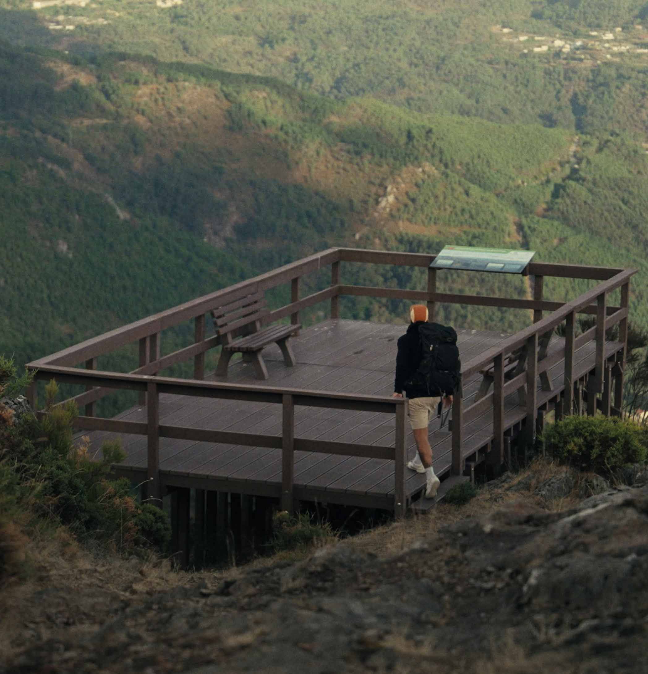 Persona caminando que entra en un mirador de plástico reciclado marrón Floema en un espacio natural. Se ve una montaña cubierta de vegetación.